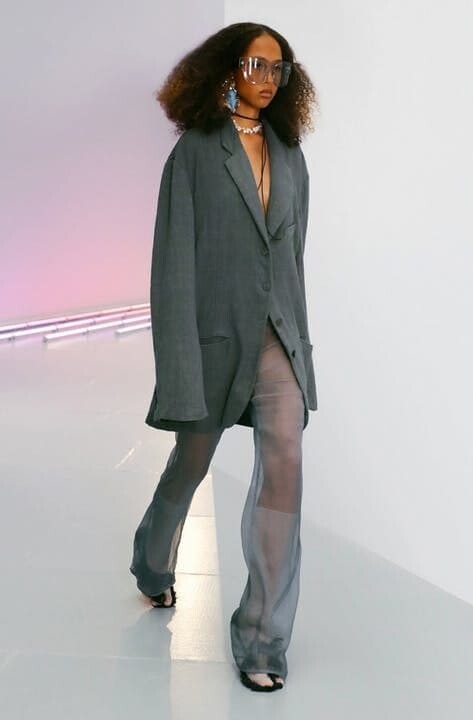 Model on the runway with a oversize gray jacket and transparent pants on a pink neon light
